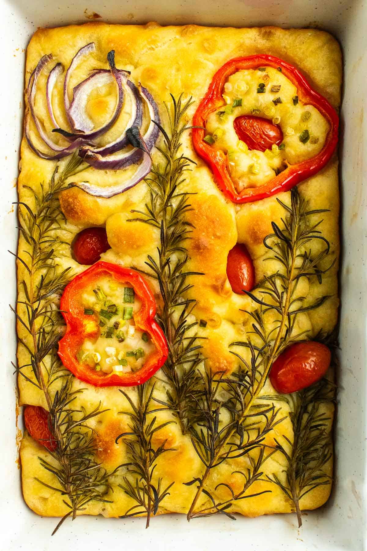 A loaf of baked focaccia bread decorated with roasted red onion, bell pepper, tomatoes, and rosemary.