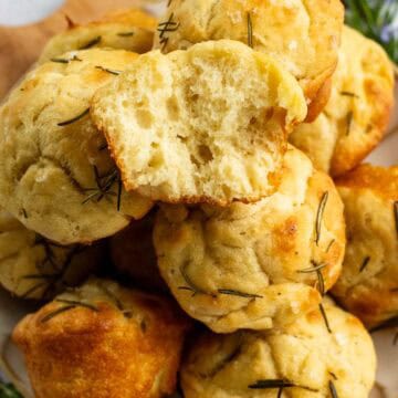 Half a dozen focaccia muffins stacked on a plate with one torn in half.