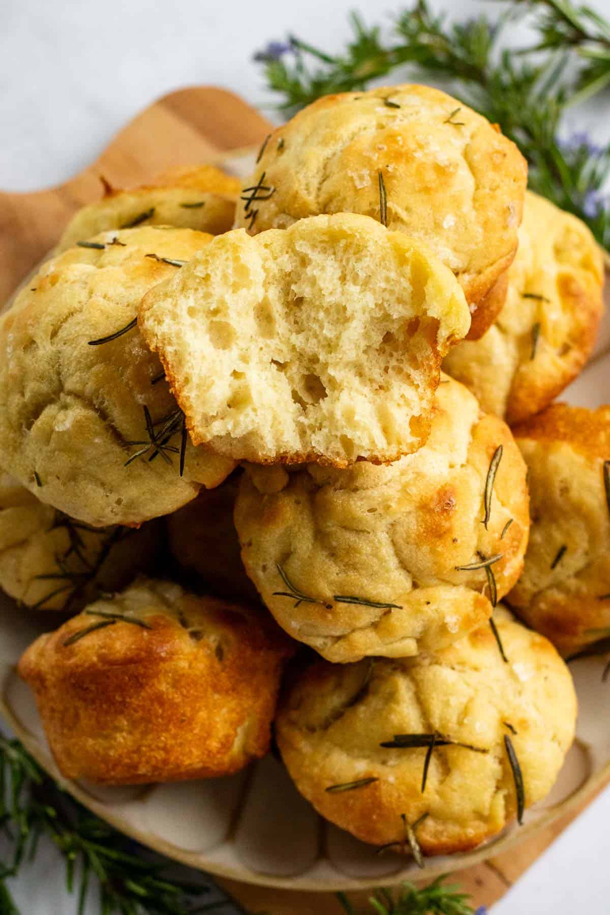 Half a dozen focaccia muffins stacked on a plate with one torn in half.