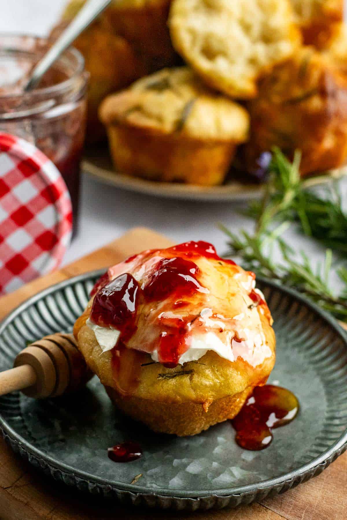 A focaccia muffin on a metal plate topped with cream cheese, red jam, and honey.