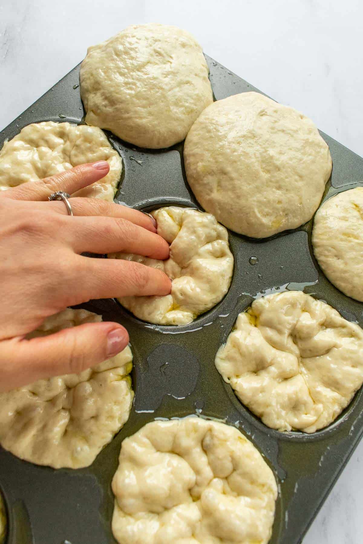 A hand pressing indentations into focaccia dough in a muffin tin.
