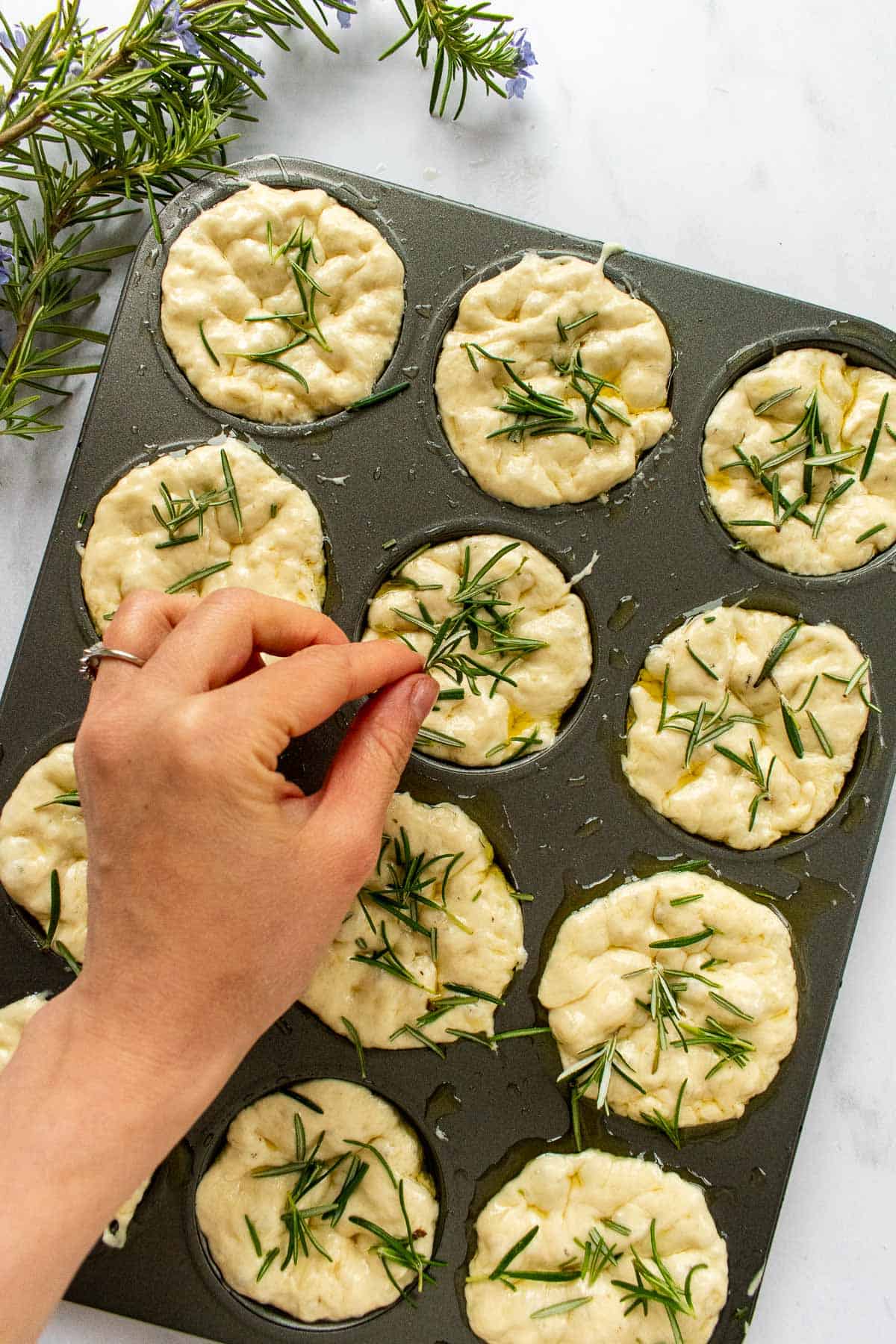A hand sprinkling fresh rosemary onto unbaked muffins in a muffin tin.