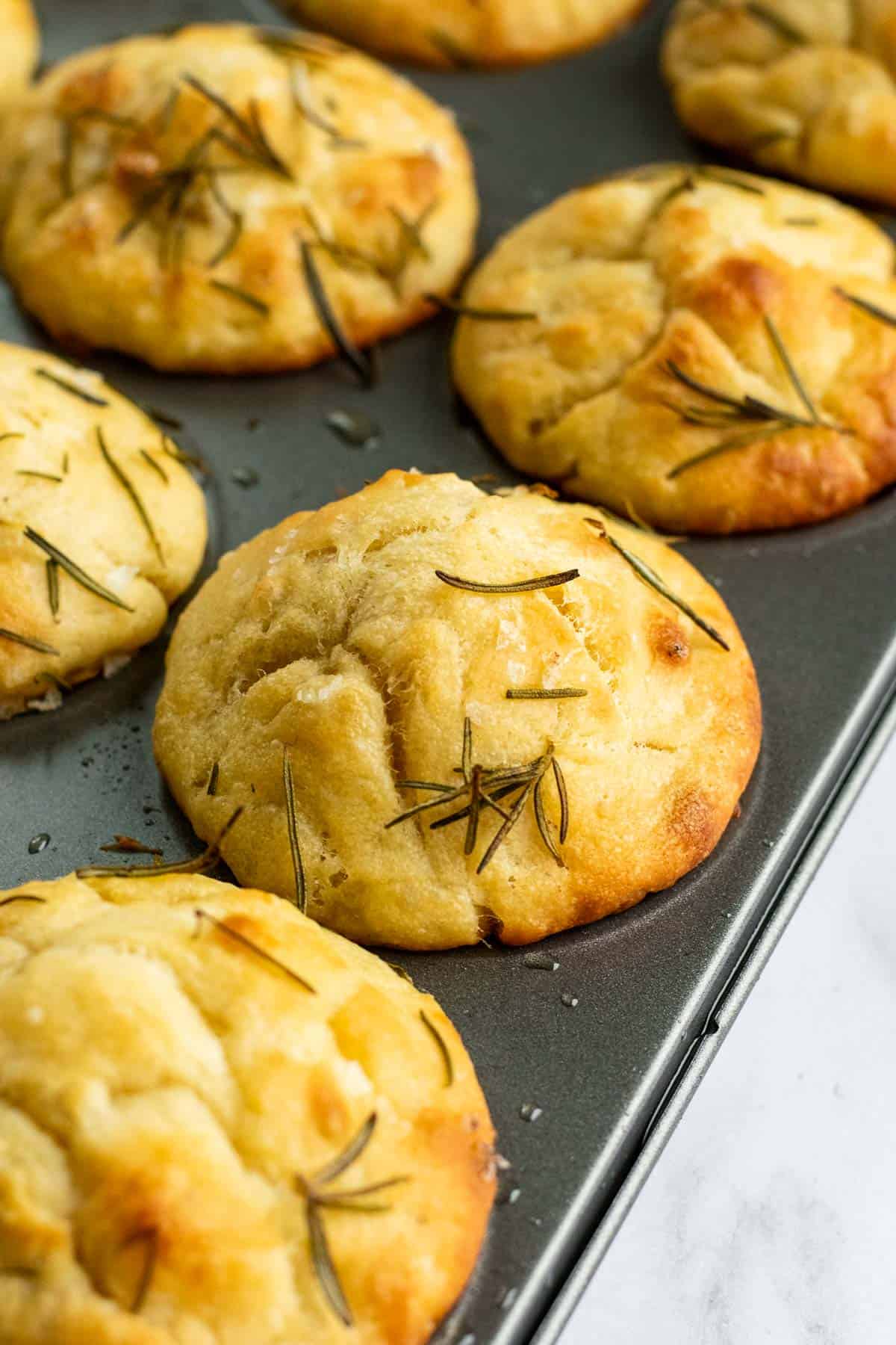 Golden baked muffins in a metal muffin pan topped with rosemary and flaky salt.