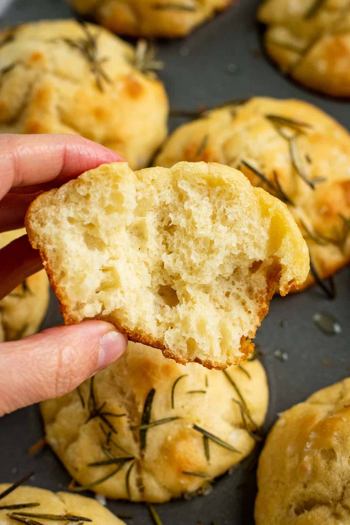 A hand holding a half-eaten focaccia muffin above a pan of more muffins.