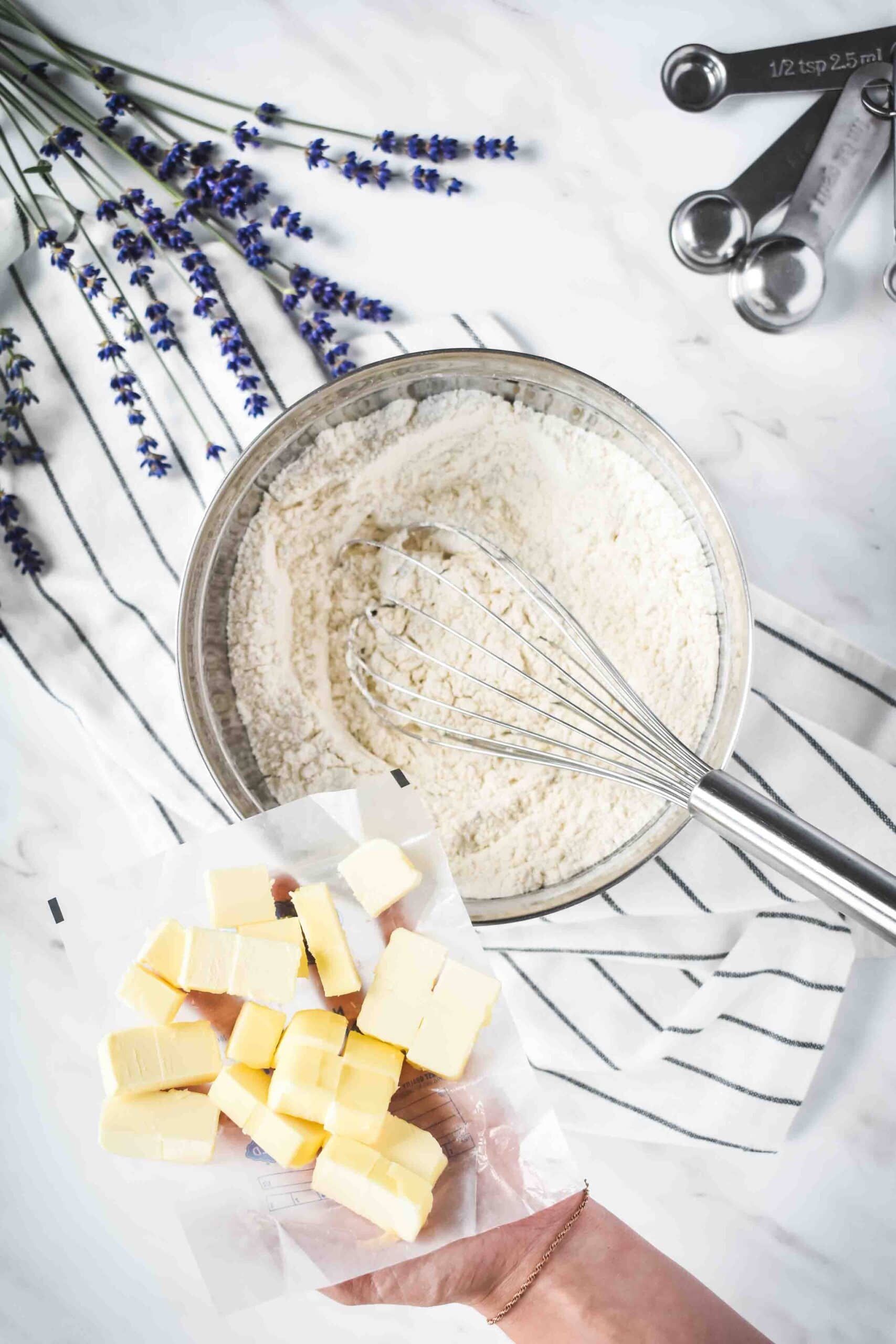 A bowl filled with flour, sugar, and salt and a hand holding cubed butter over it.