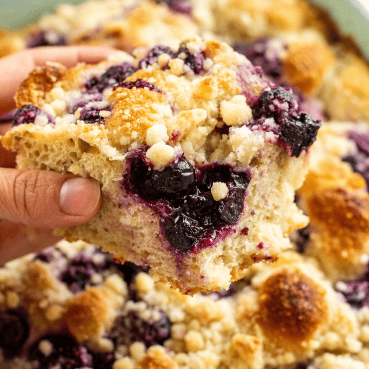 A hand holding a large square of focaccia topped with cooked blueberries and lemon streusel over a dish of more bread.
