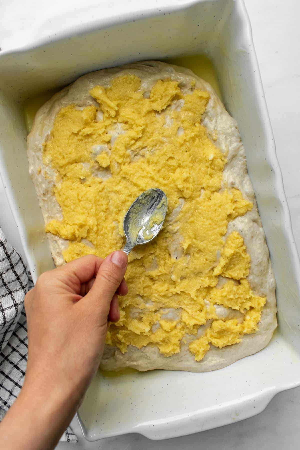 A hand spooning creamy almond frangipane on bread dough in a white baking dish.