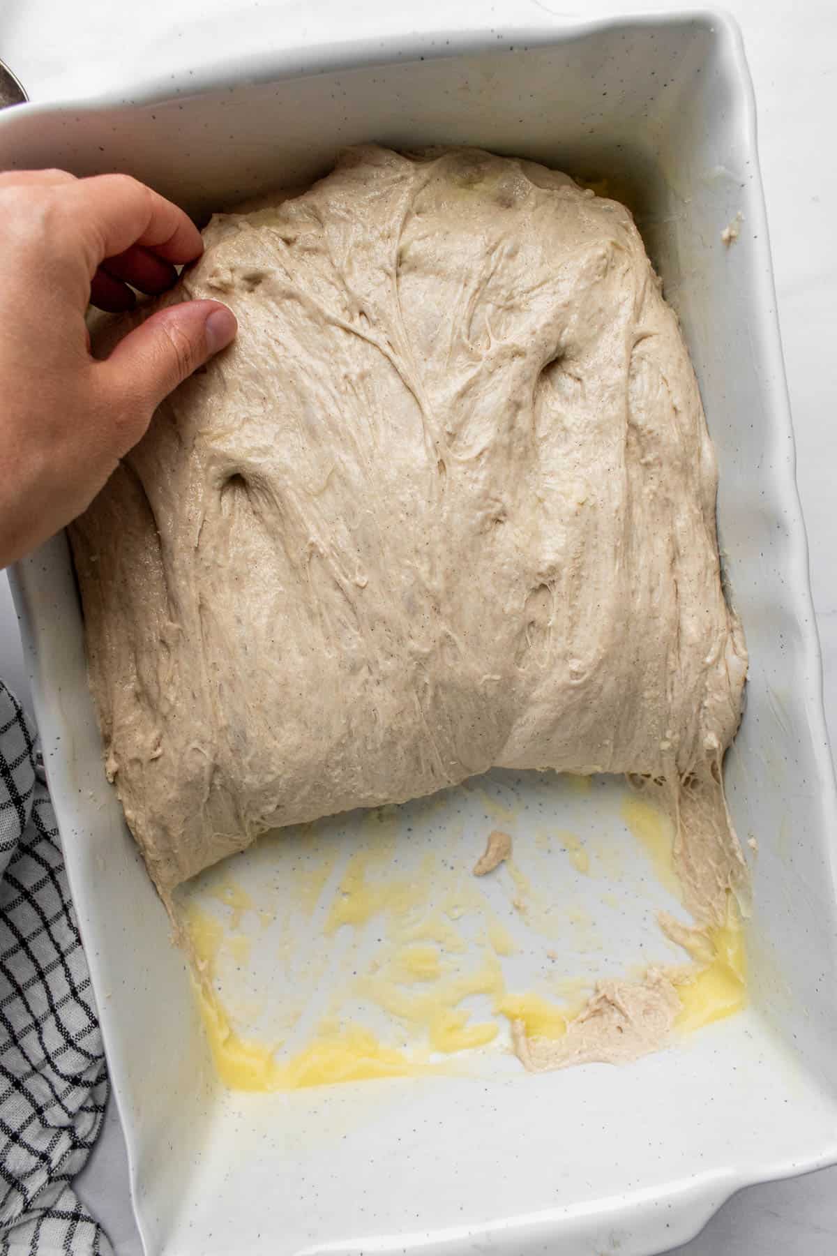A hand folding focaccia dough in half in a white baking dish.