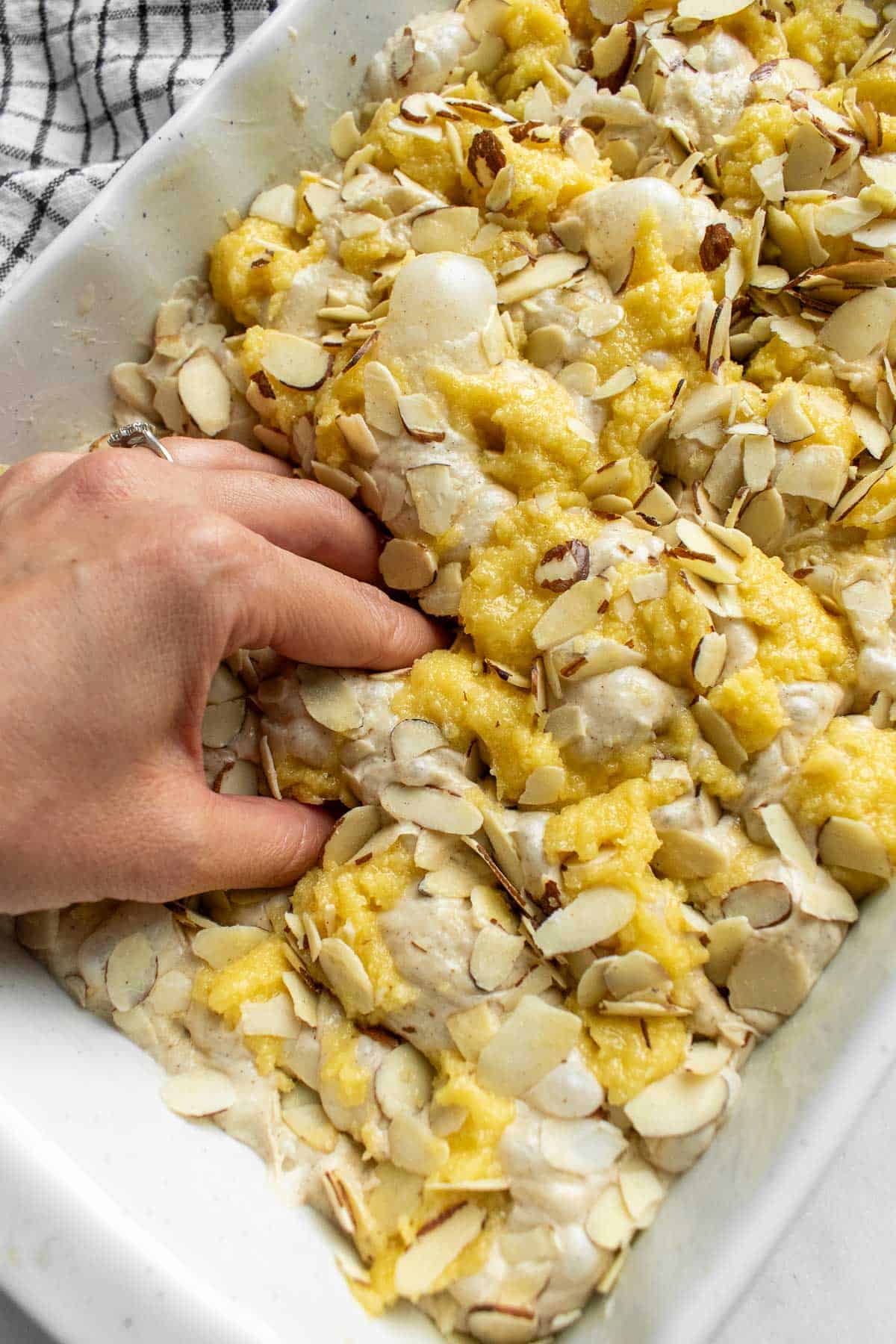 A hand dimpling almond croissant focaccia dough in a large white baking dish topped with sliced almonds.