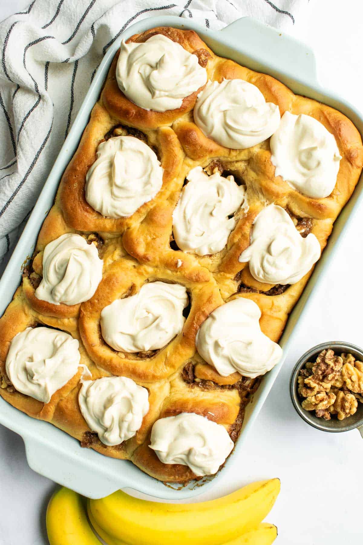 Twelve golden cinnamon rolls in a baking dish topped with dollops of whipped cream next to bananas and walnuts.