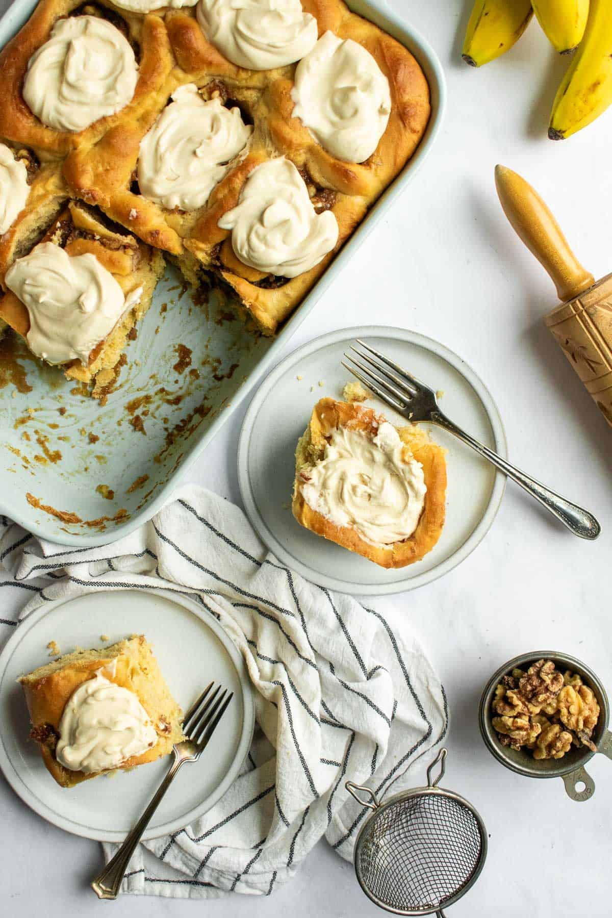 Two cinnamon rolls topped with whipped cream on grey plates next to more cinnamon rolls in a baking dish.