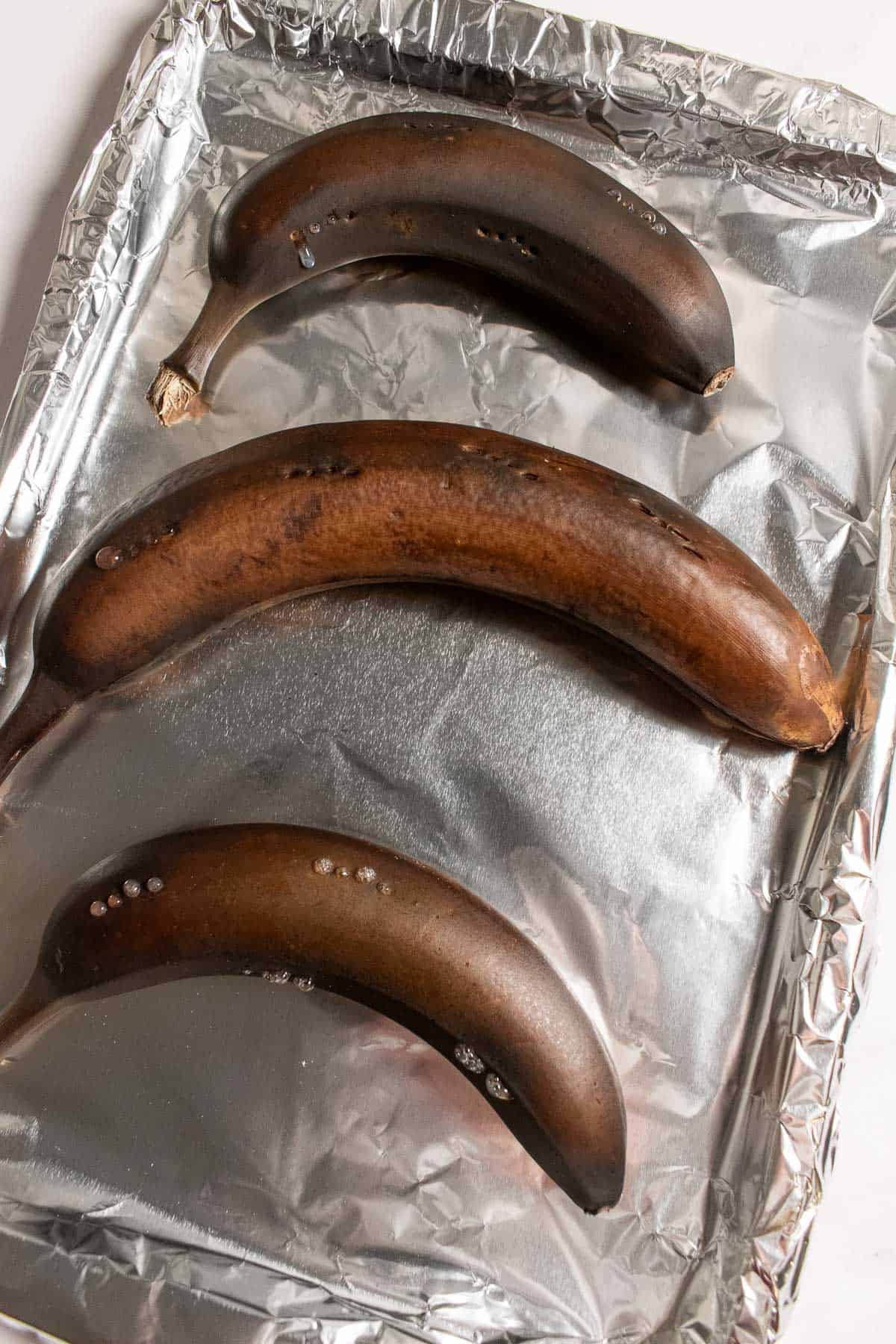 Three black, roasted bananas on a baking sheet lined with aluminum foil.