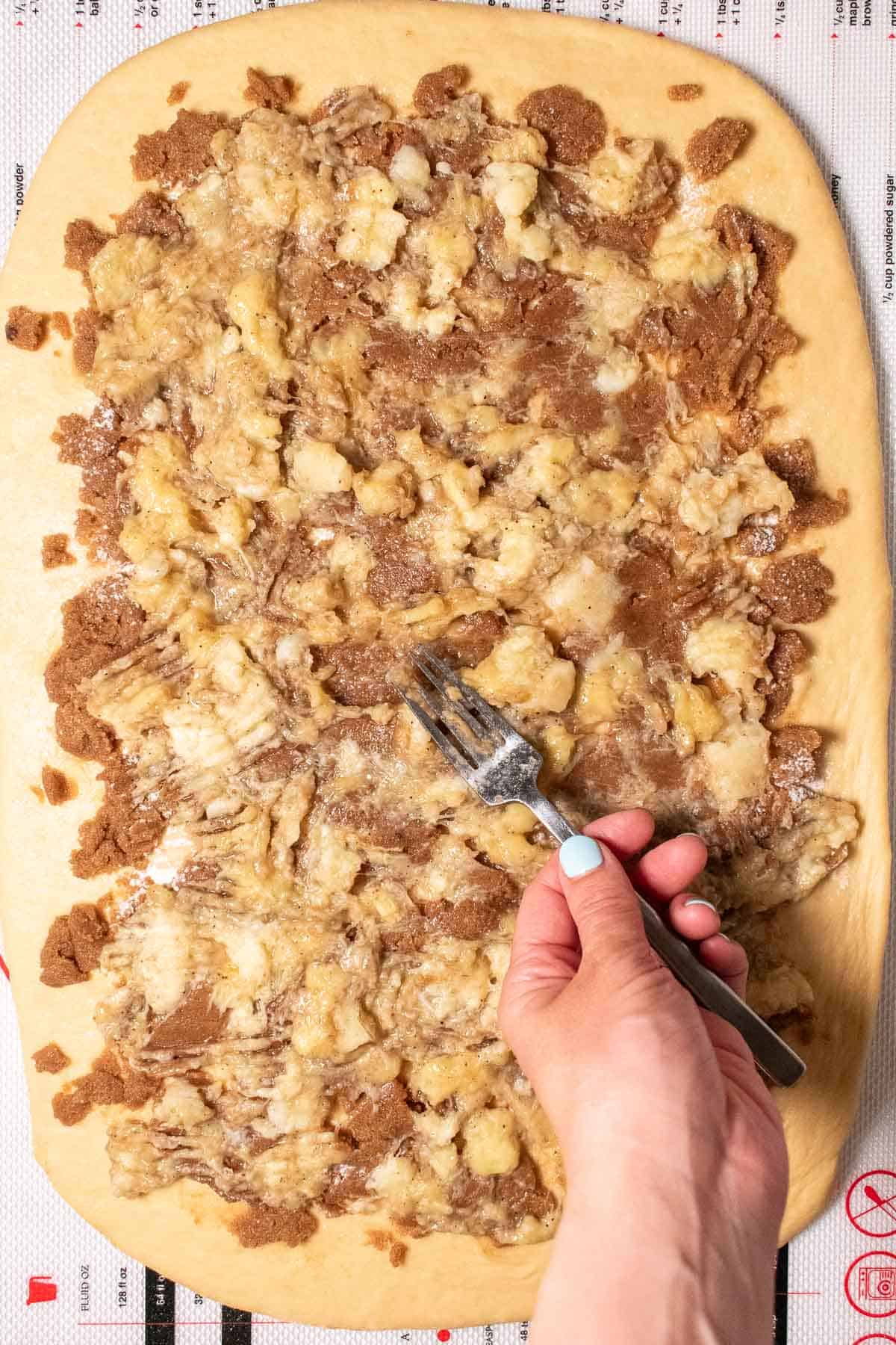A hand spreading mashed bananas onto cinnamon roll dough with a fork.