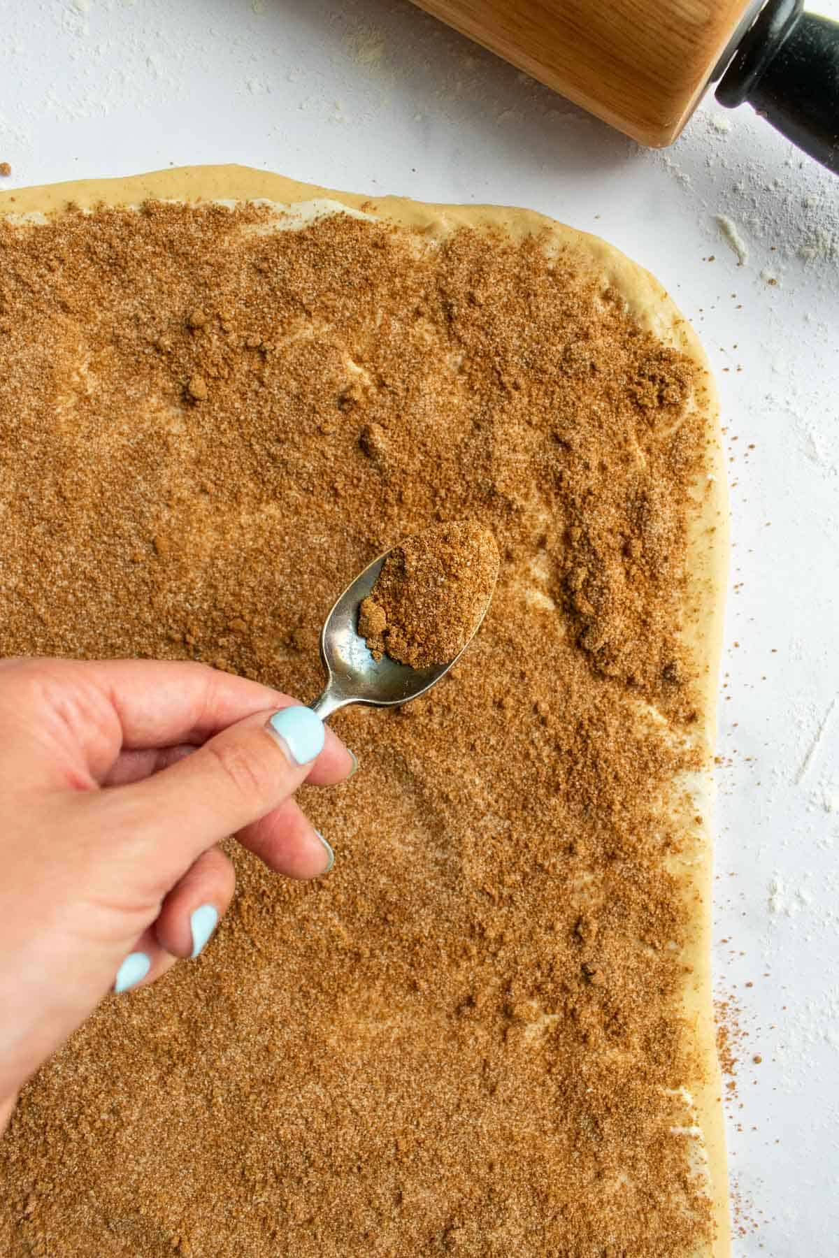 A hand sprinkling cinnamon sugar from a metal spoon onto rolled-out dough.