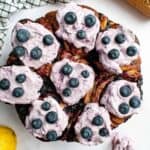 Baked cinnamon rolls in a round cake pan topped with purple whipped cream and fresh blueberries.