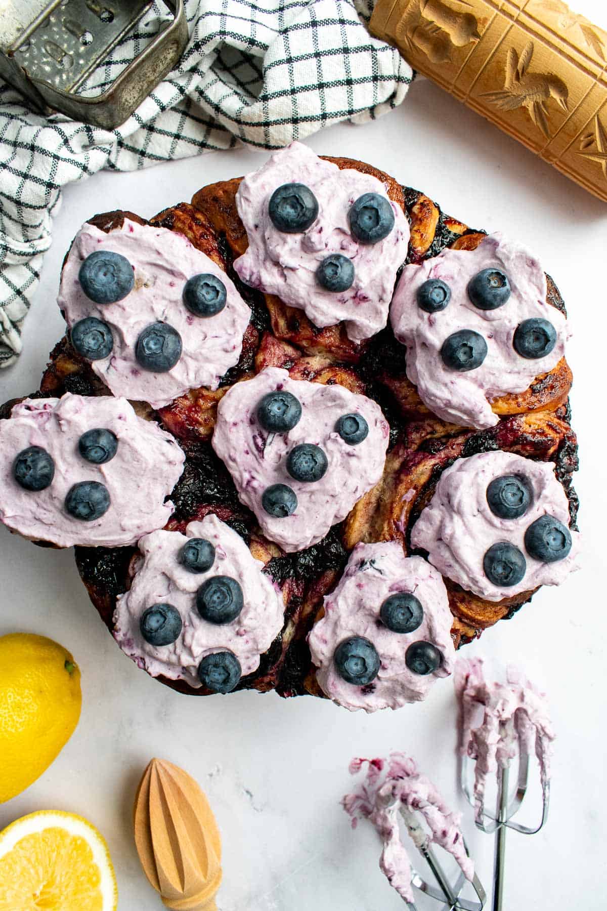 Eight cinnamon rolls in a round pan topped with purple whipped cream and blueberries.