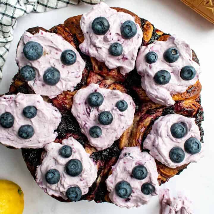 Baked cinnamon rolls in a round cake pan topped with purple whipped cream and fresh blueberries.