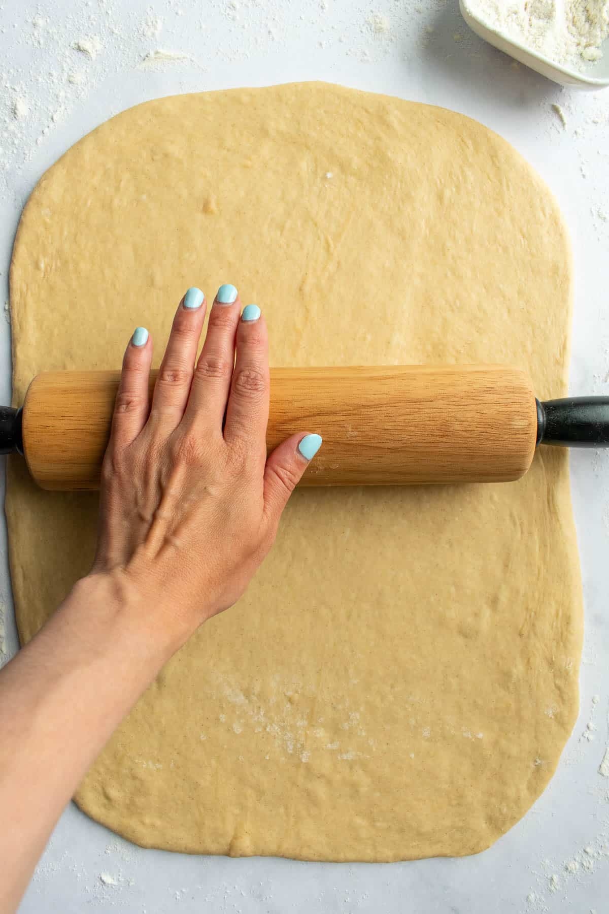 A hand rolling out cinnamon roll dough on a light surface with a wooden rolling pin.