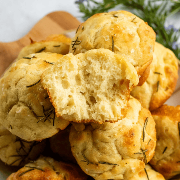 A dozen focaccia muffins piled on a cutting board with the top muffin torn in half.