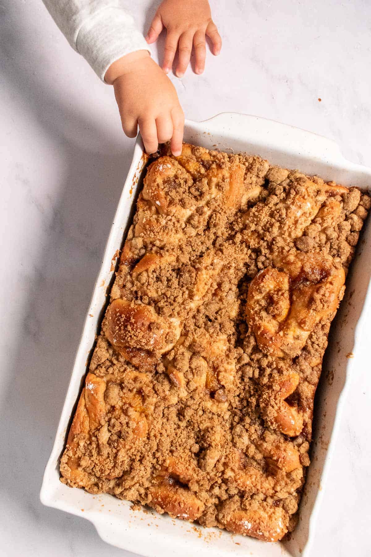 A dozen baked cinnamon rolls topped with crumbly streusel in a white baking dish as hands try to reach for a cinnamon roll.