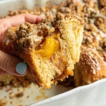 A hand holding a large cinnamon roll with peaches inside and crumbly streusel and white glaze on top.