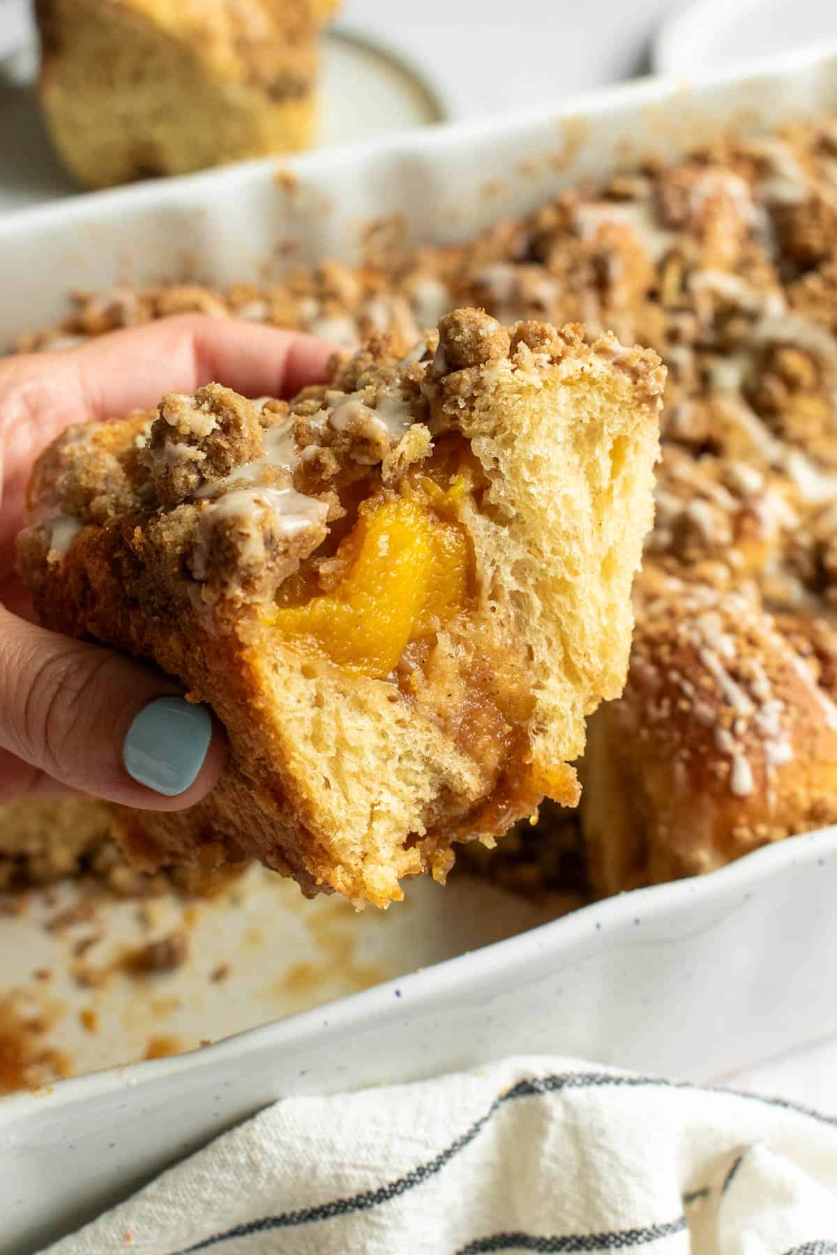 A hand holding a large cinnamon roll with peaches inside and crumbly streusel and white glaze on top.
