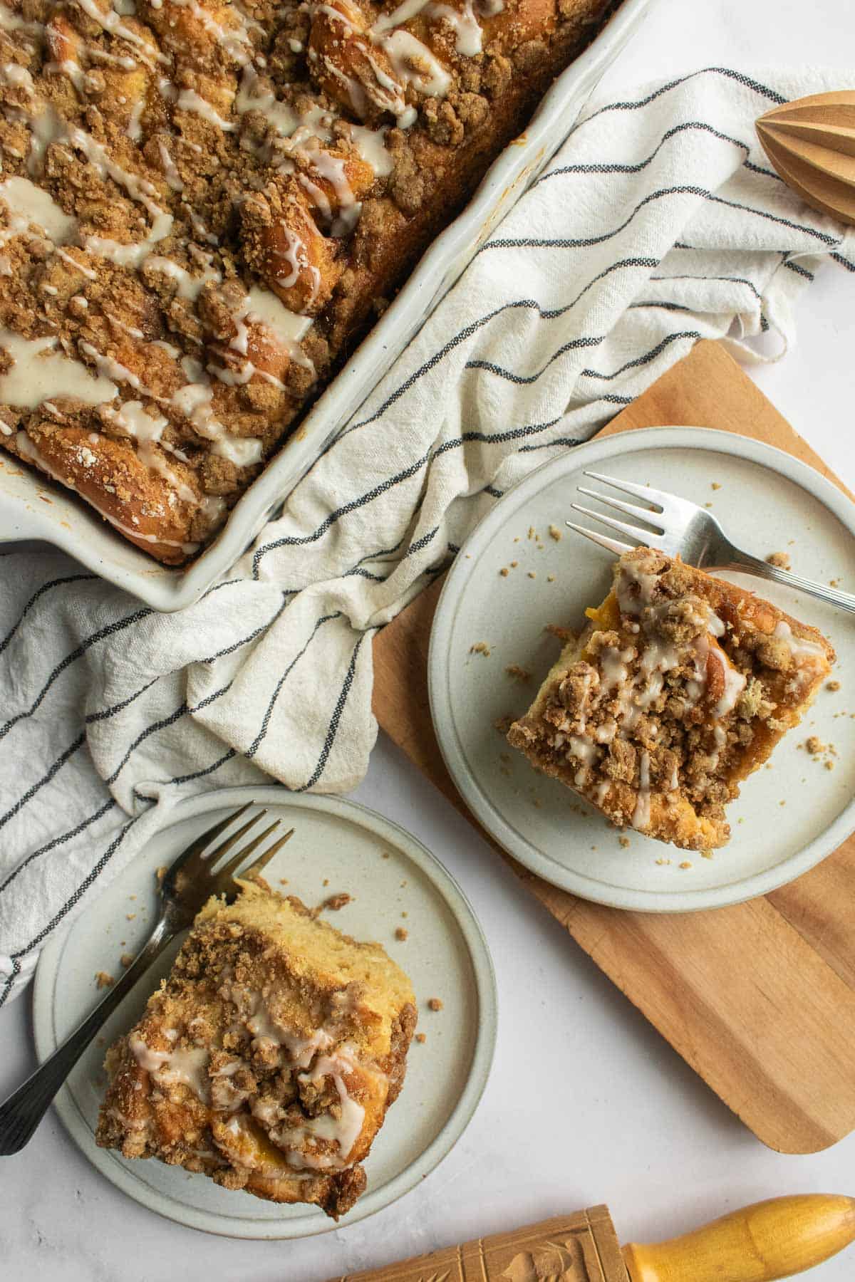Two cinnamon rolls on grey plates topped with crumbly streusel and white glaze with metal forks next to them.