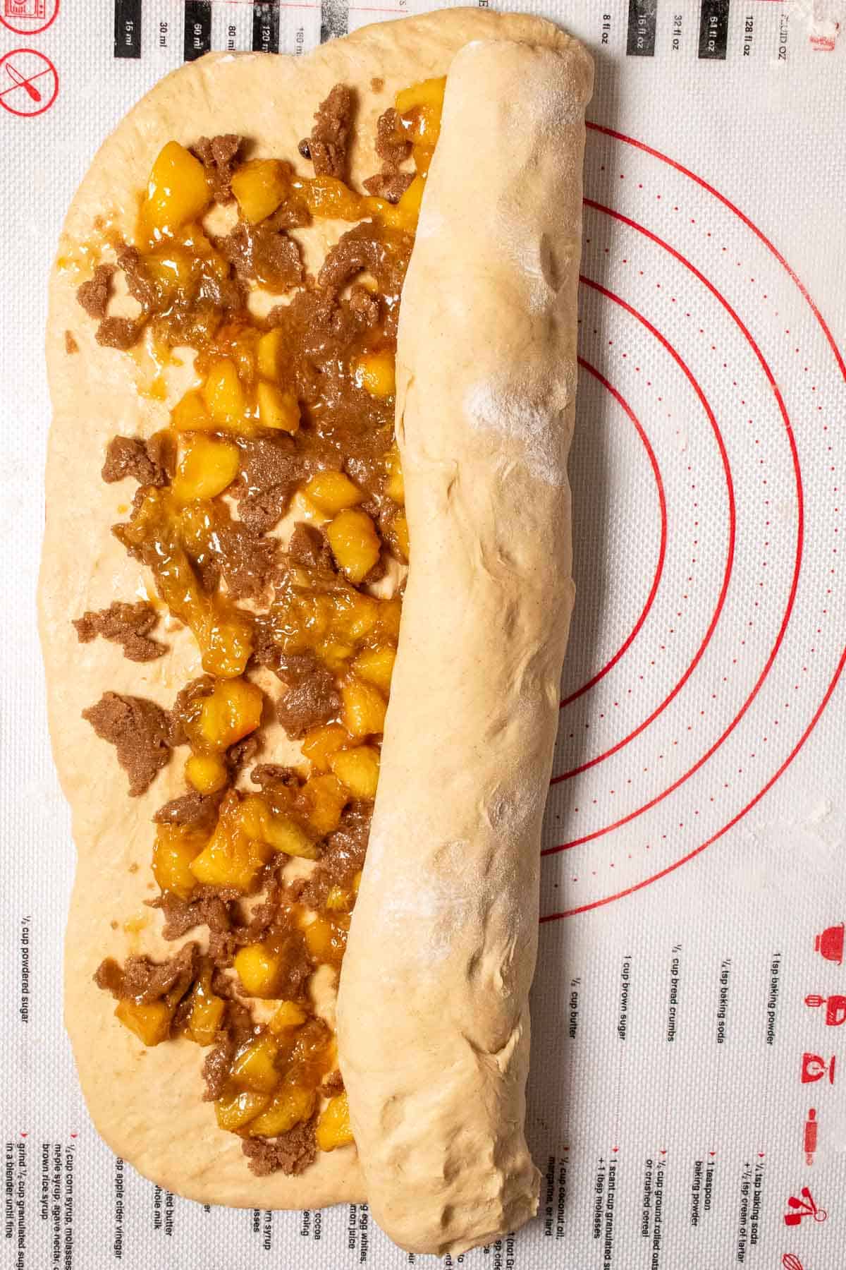 Brown sugar, butter, and cooked peaches spread on cinnamon roll bread dough being rolled up into a log.