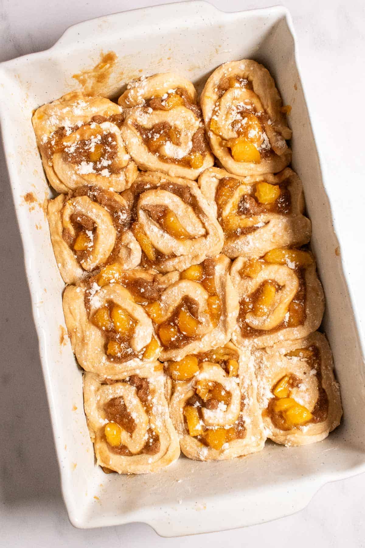 Twelve cinnamon rolls filled with brown sugar and cooked peaches in a white baking dish.