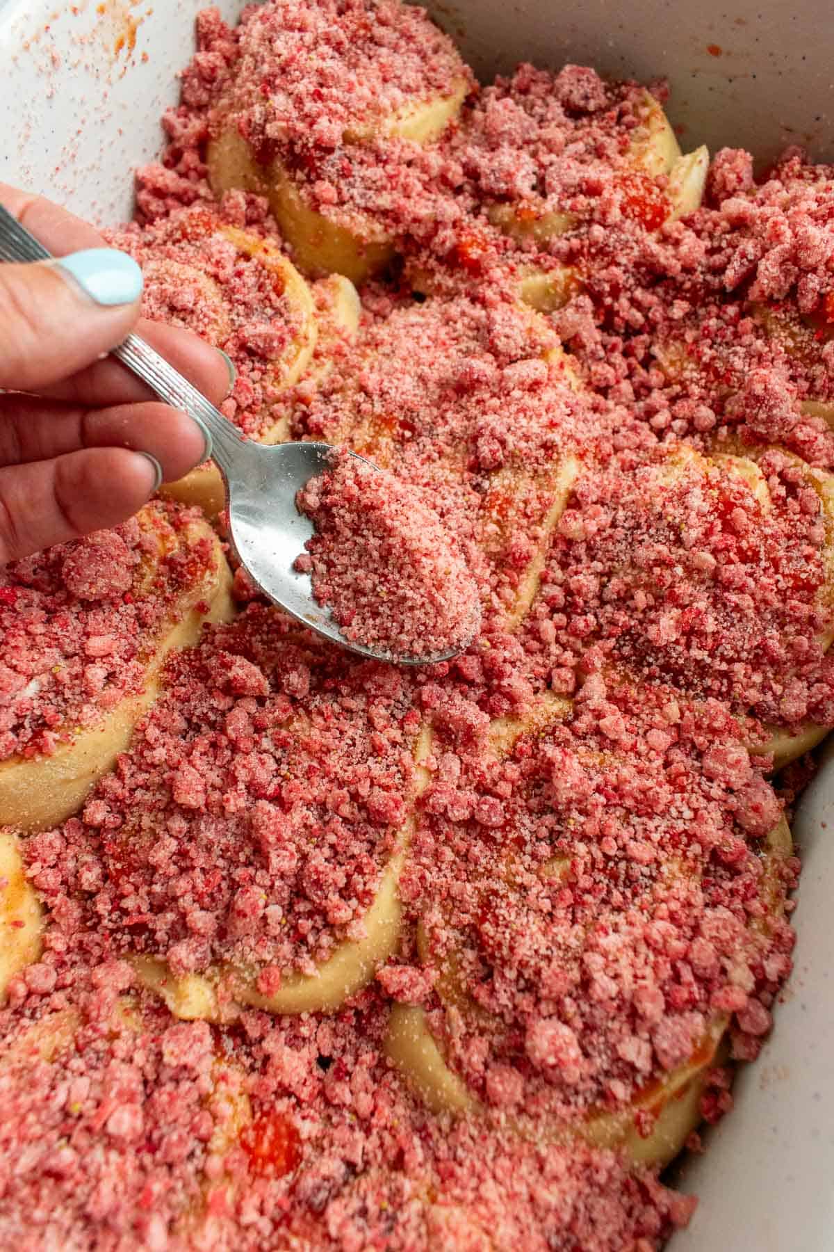 A hand sprinkling pink strawberry streusel onto unbaked cinnamon rolls.