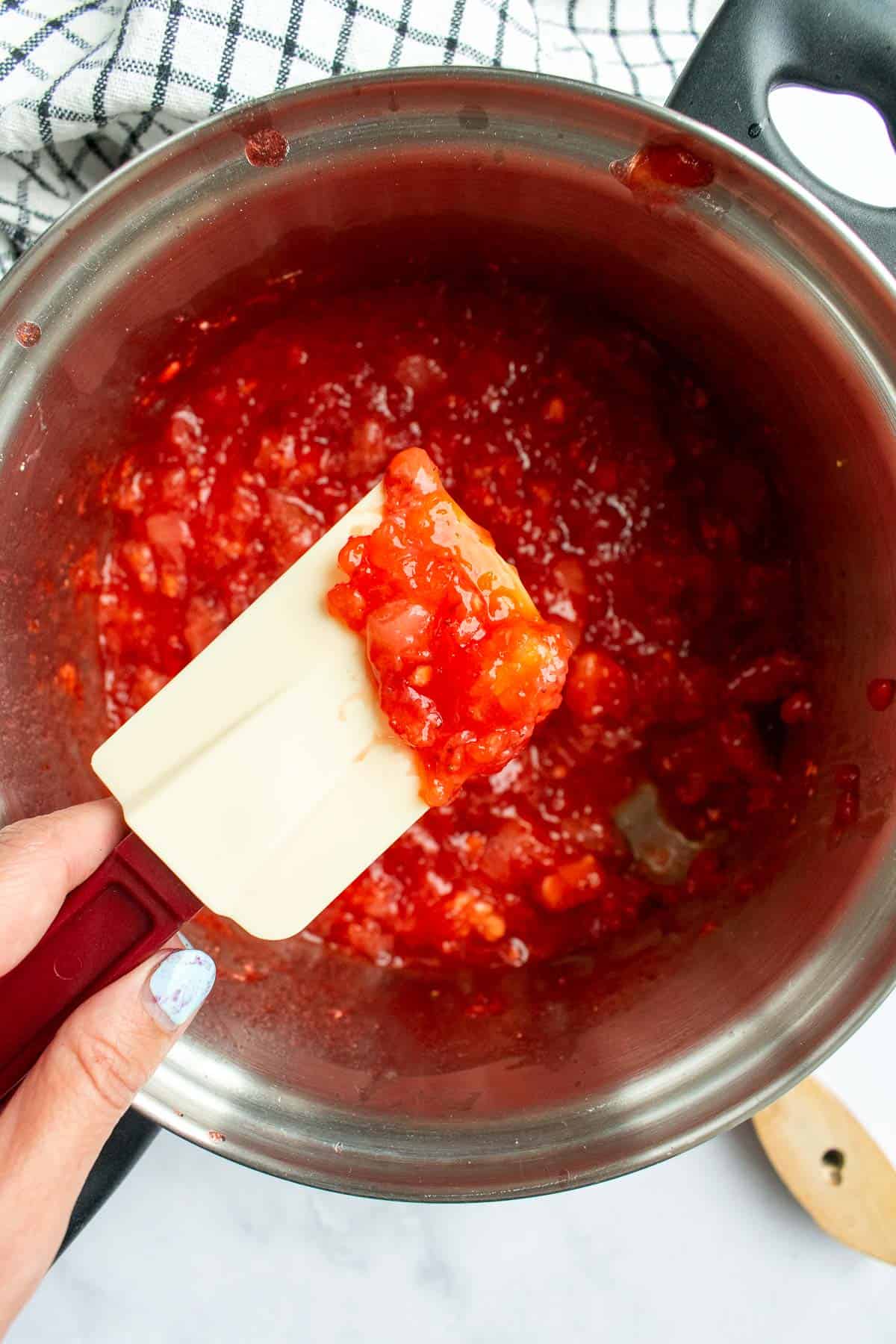 A hand holding a spatula covered in thick, red jam over a saucepan with more jam.
