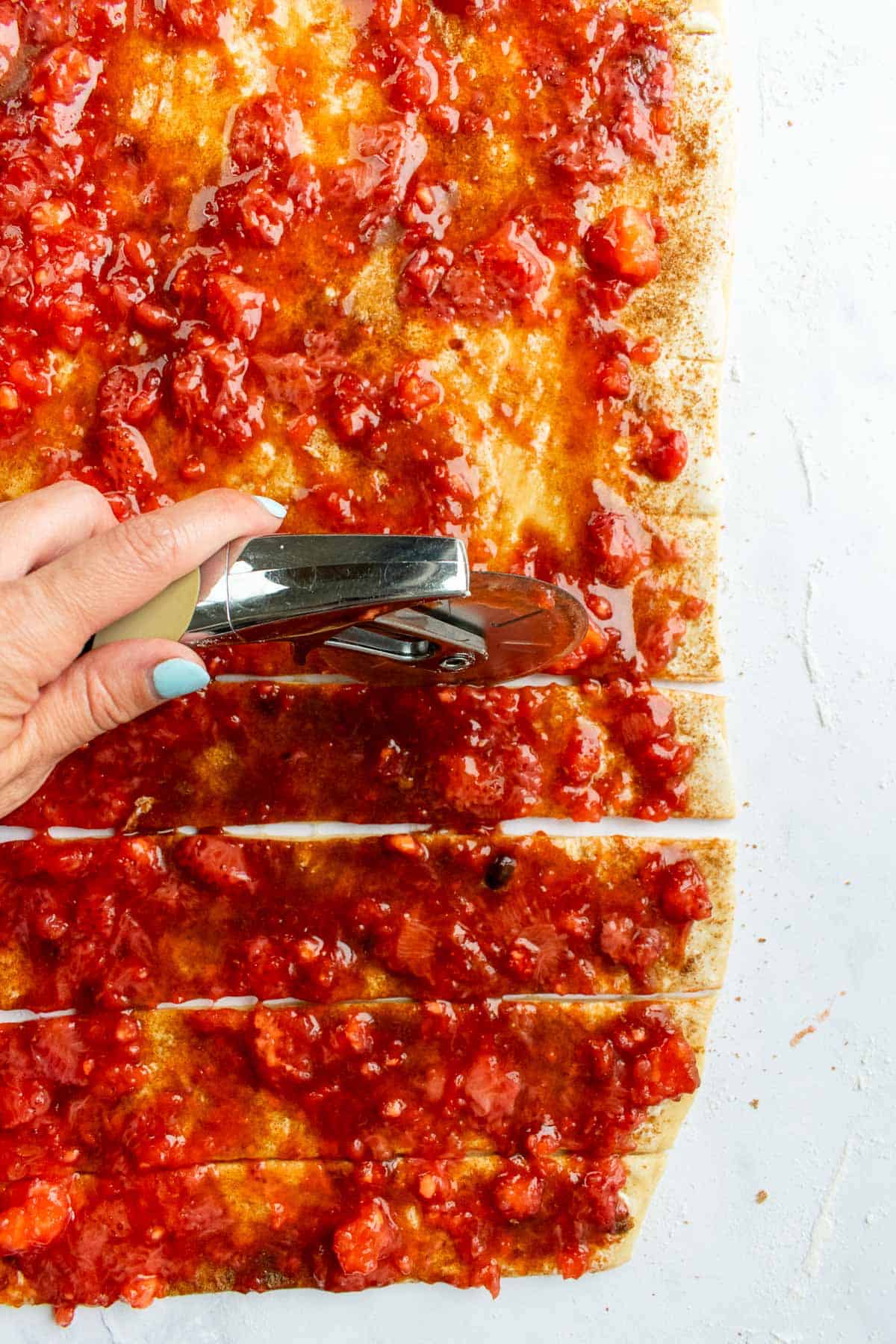 A hand cutting dough covered in strawberry jam into strips with a pizza cutter.