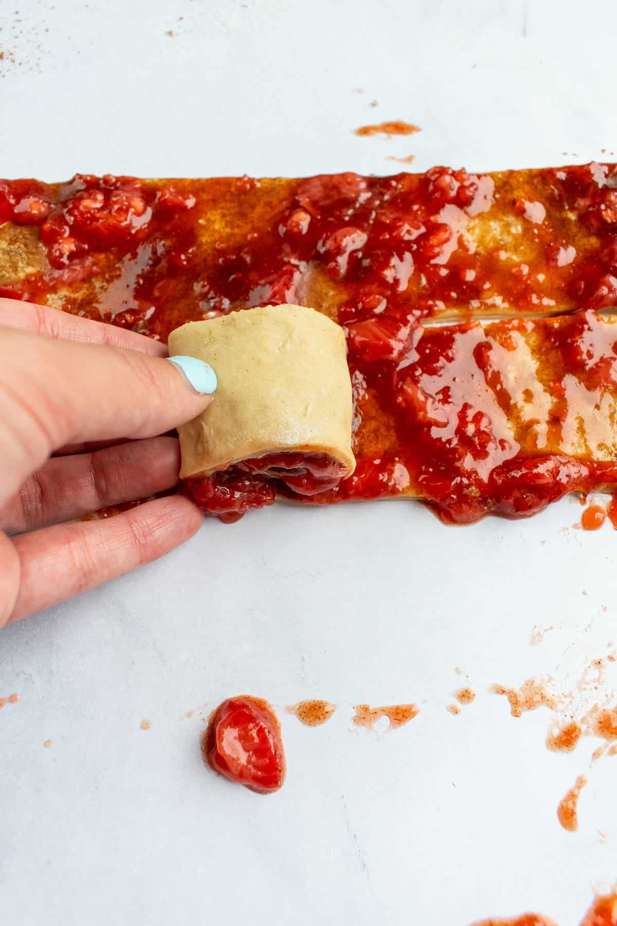 A hand rolling strawberry jam into cinnamon roll dough on a light surface.