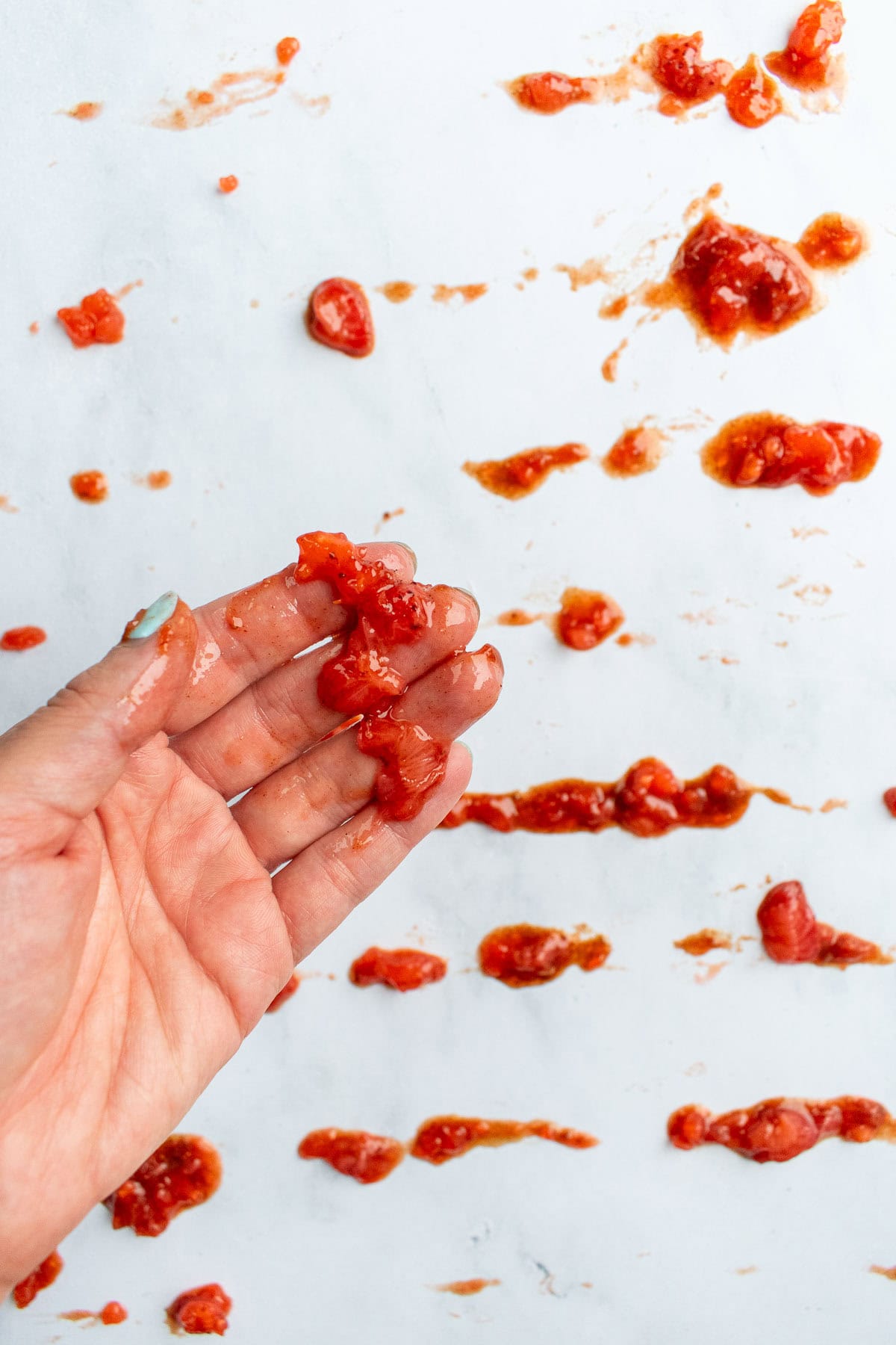 A hand holding cooked strawberries over a counter covered in chunks of strawberry jam.