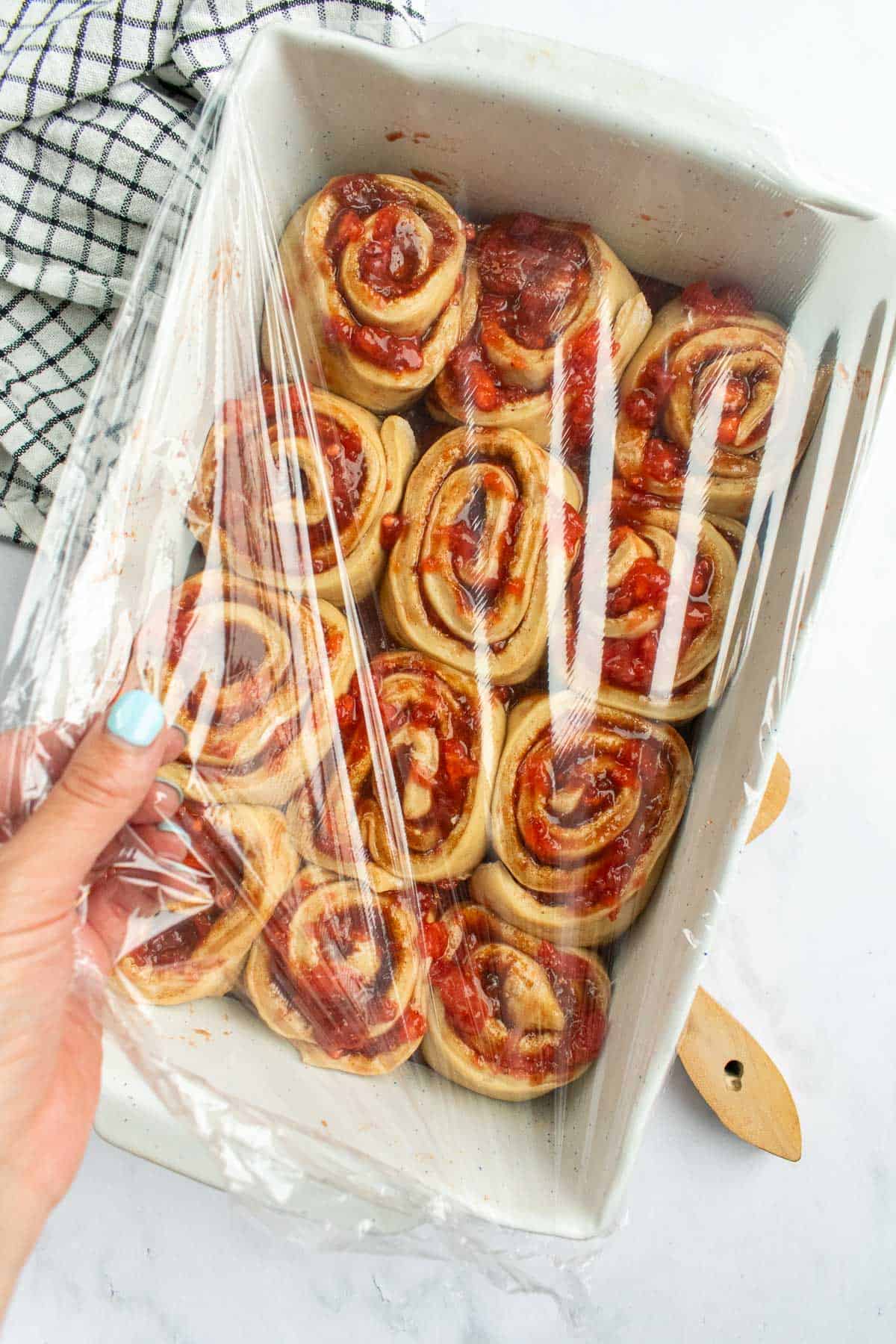 A hand holding plastic wrap over a baking dish of strawberry cinnamon rolls.
