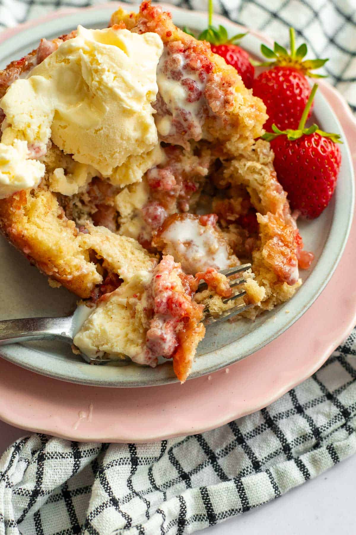 A half-eaten strawberry cinnamon roll topped with melting vanilla ice cream on a plate next to fresh strawberries.