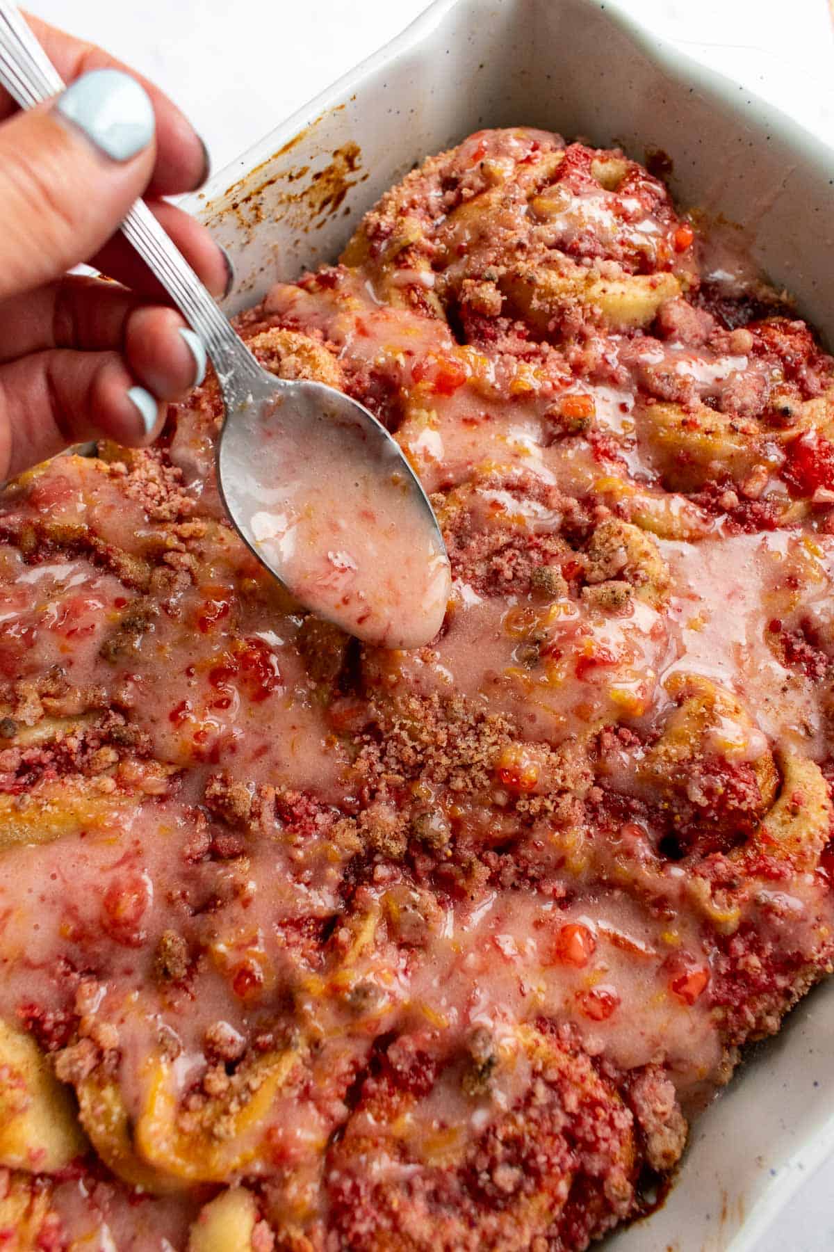 A hand drizzling pink strawberry glaze onto baked cinnamon rolls topped with crumbly streusel.