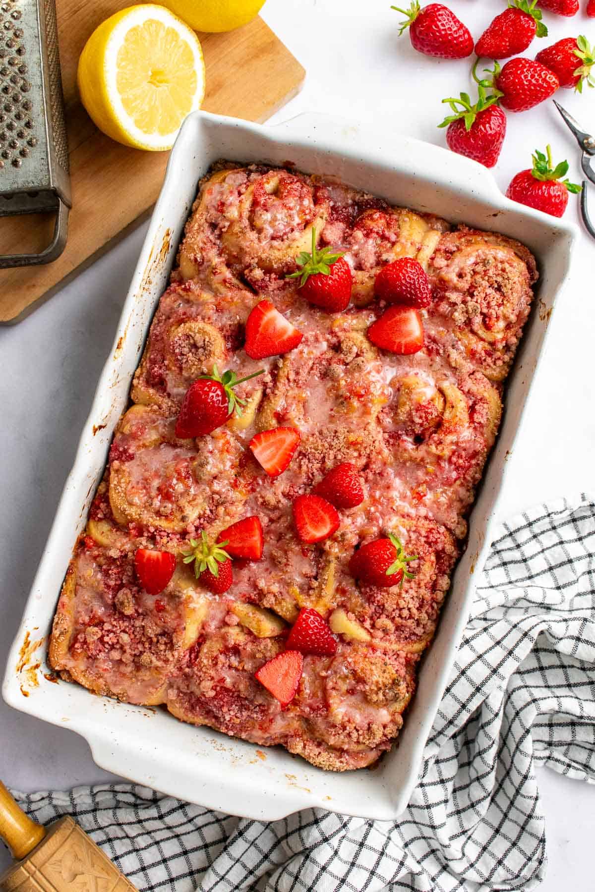 A white baking dish filled with a dozen cinnamon rolls topped with pink streusel, pink glaze, and fresh strawberries.