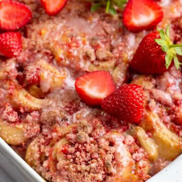 Cinnamon rolls in a white baking dish topped with crumbly pink streusel and fresh strawberries.