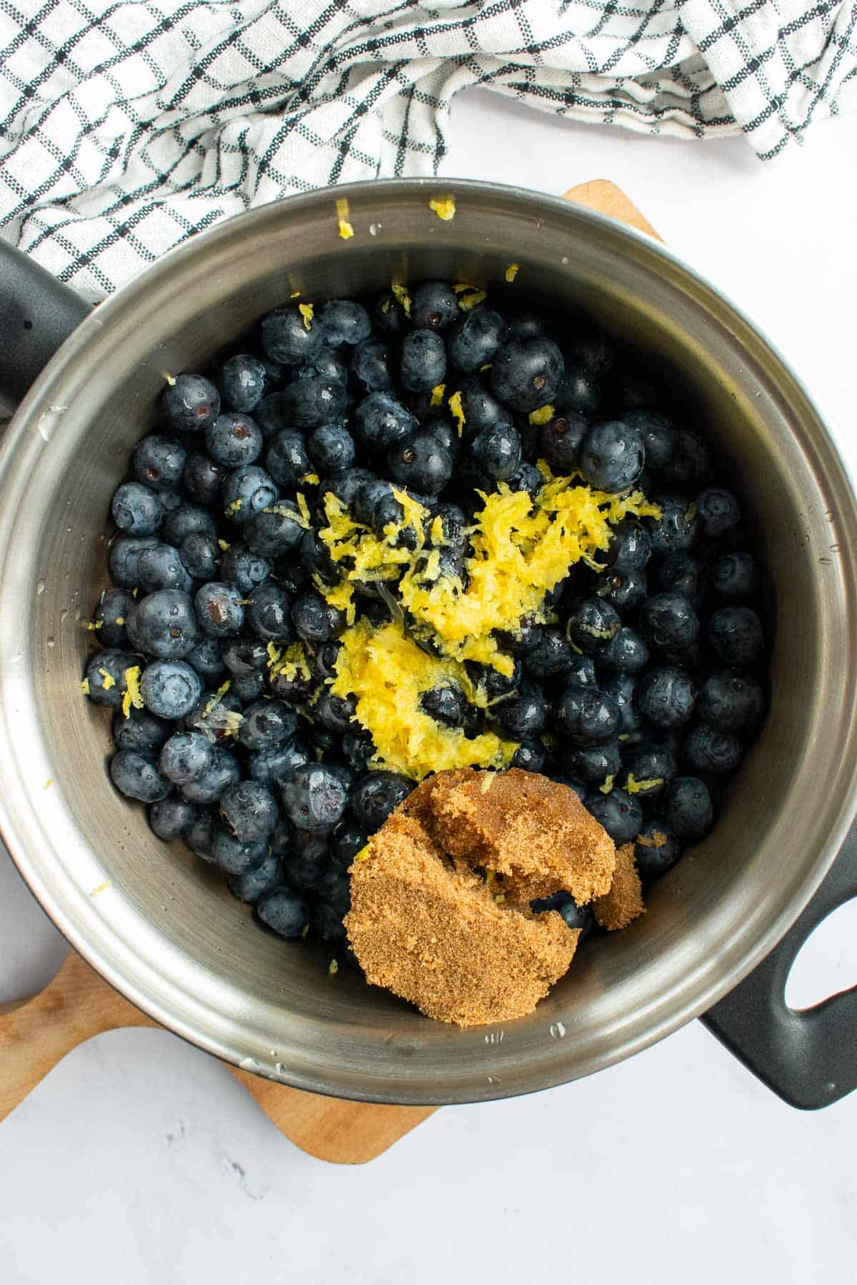 A metal saucepan with fresh blueberries, lemon zest, and brown sugar on a light surface.