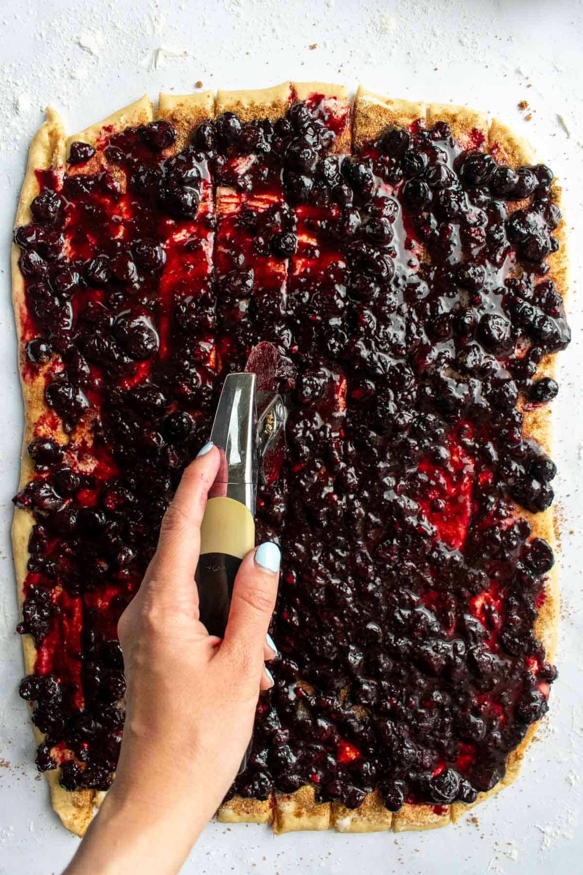 A hand cutting cinnamon roll dough covered in blueberry jam with a pizza cutter into long strips.