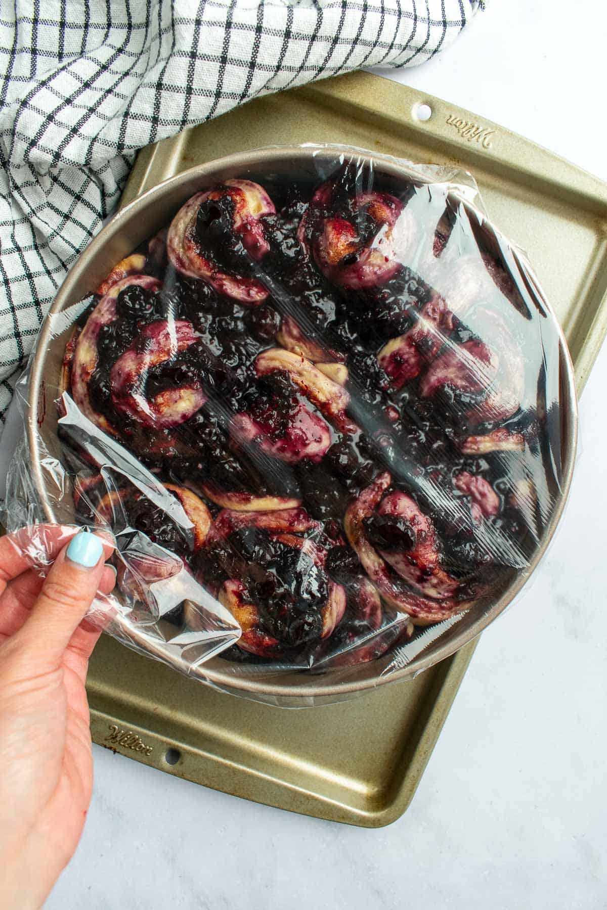 A hand pulling plastic wrap over a round cake pan filled with unbaked blueberry cinnamon rolls.