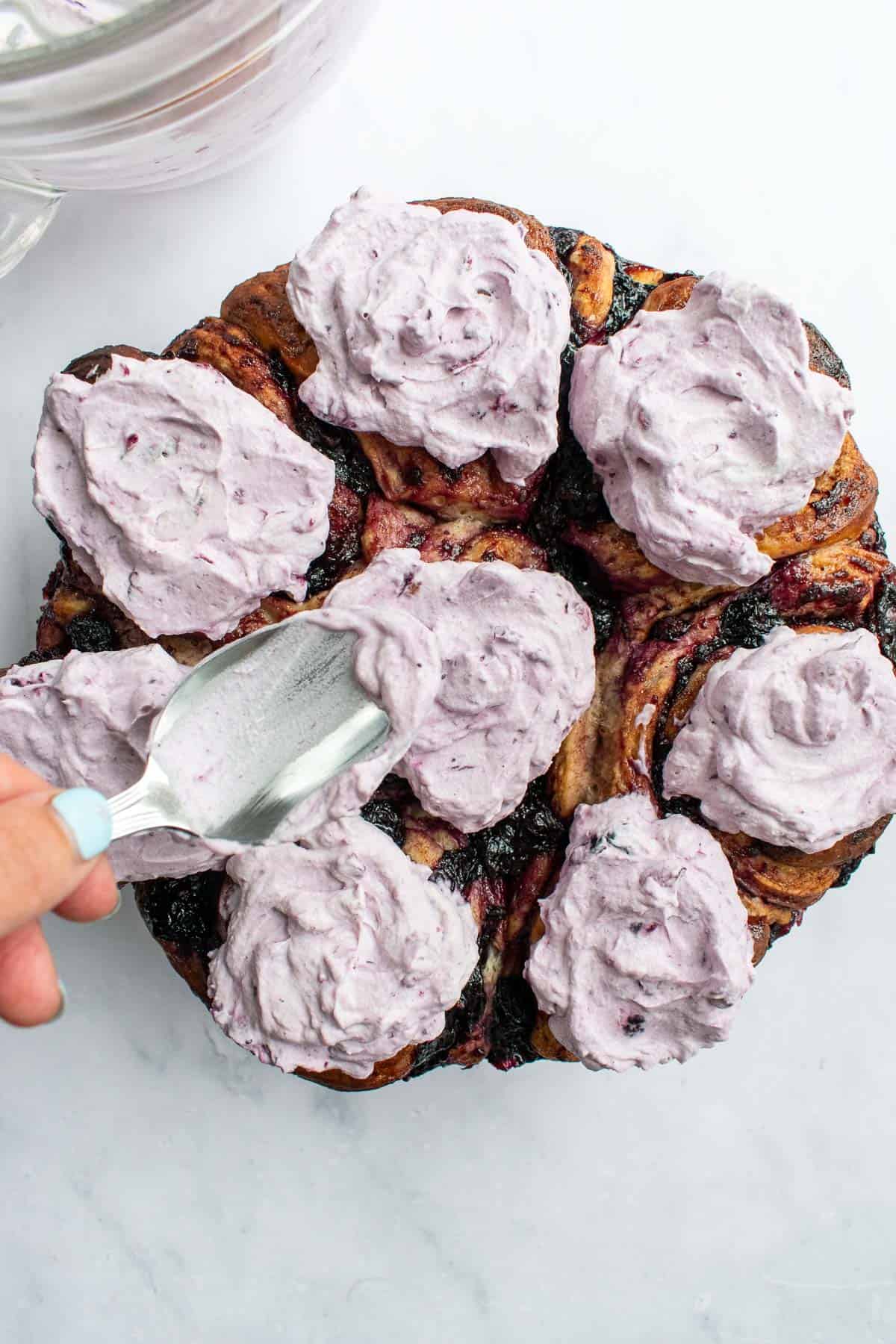 A hand dolloping light purple blueberry whipped cream onto cinnamon rolls with a metal spoon.
