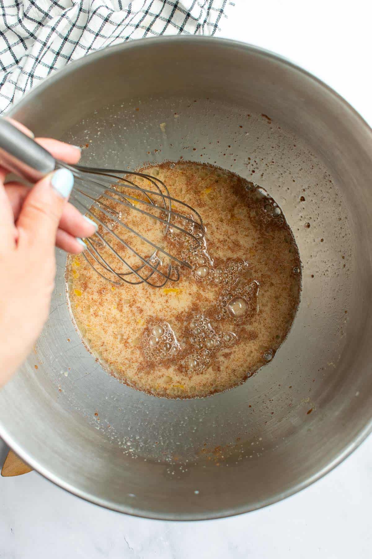 A hand whisking a light brown liquid of milk, eggs, brown sugar, and yeast in a metal bowl.
