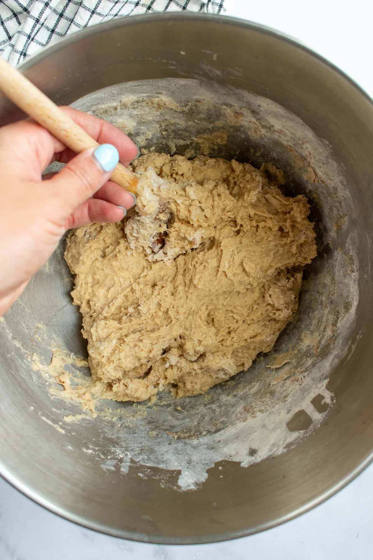 A hand mixing a sticky bread dough in a large metal bowl.