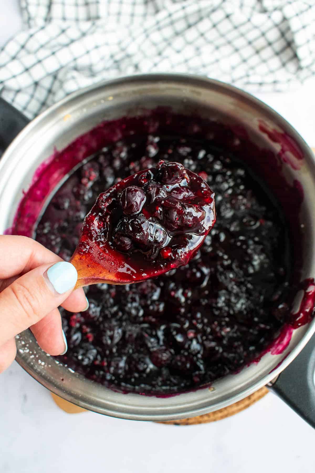 A hand holding a wooden spoon with blueberry jam on it over a saucepan with more jam inside.