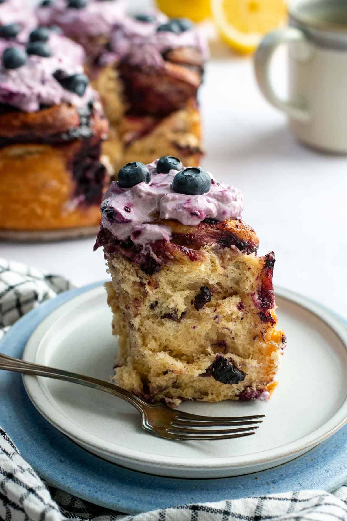 A tall blueberry cinnamon roll on a grey plate topped with light purple whipped creme fraiche and fresh blueberries.