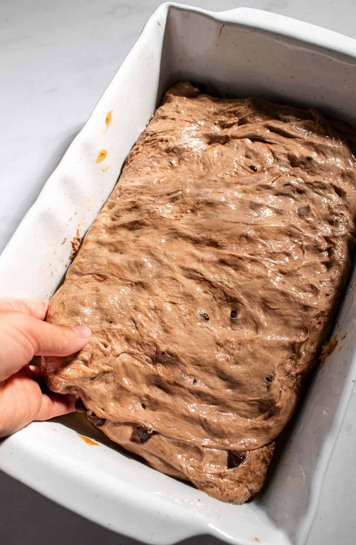 A hand spreading chocolate-filled focaccia dough in a large baking dish.