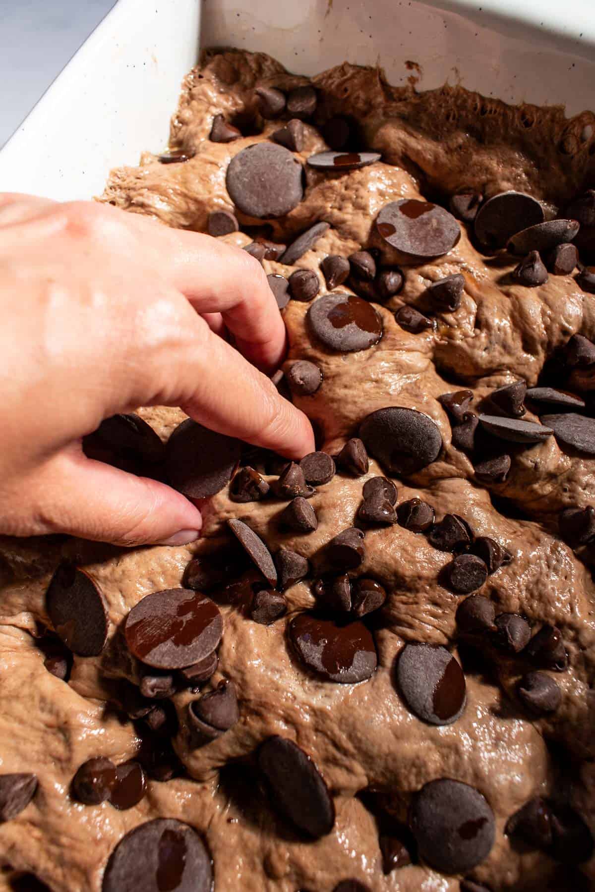 A hand dimpling brown focaccia dough in a baking dish that's topped with chocolate chips.