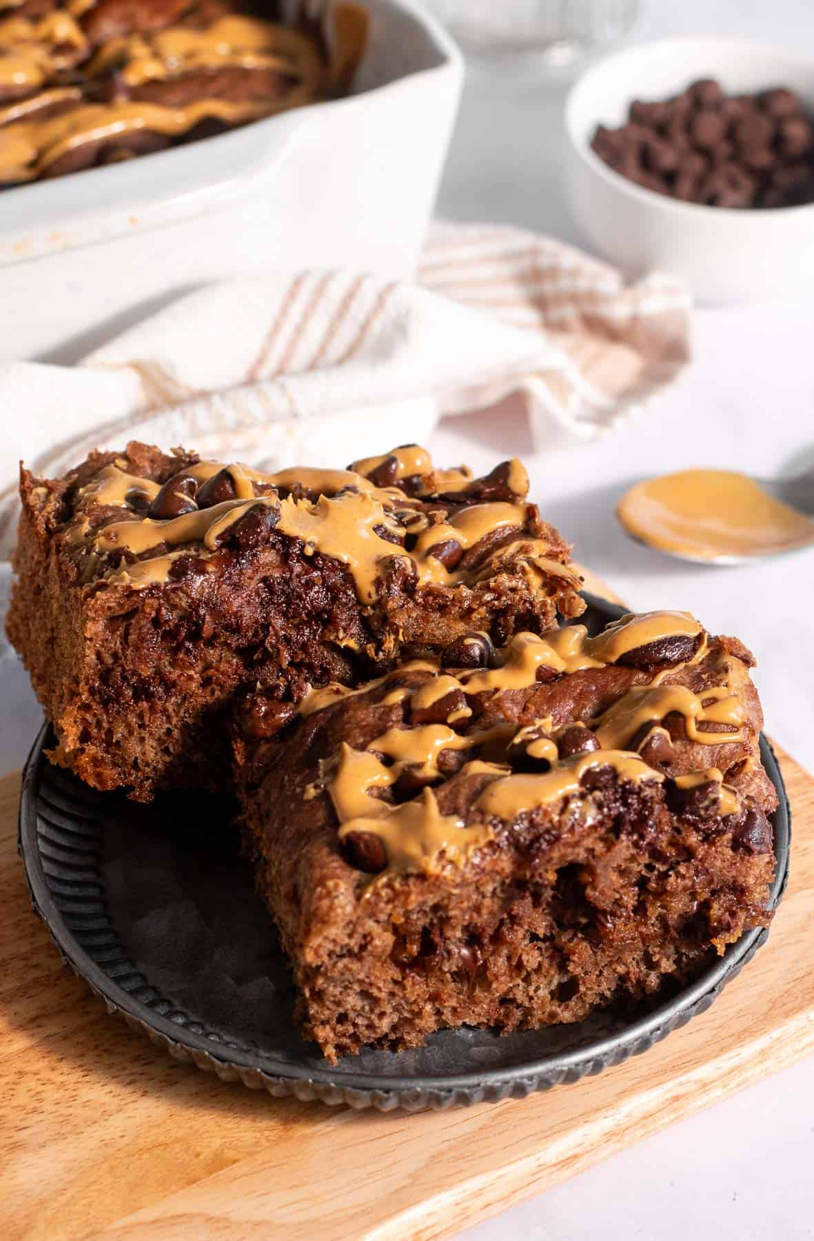 Two large squares of chocolate focaccia drizzled with peanut butter on a plate next to a bowl of chocolate chips.